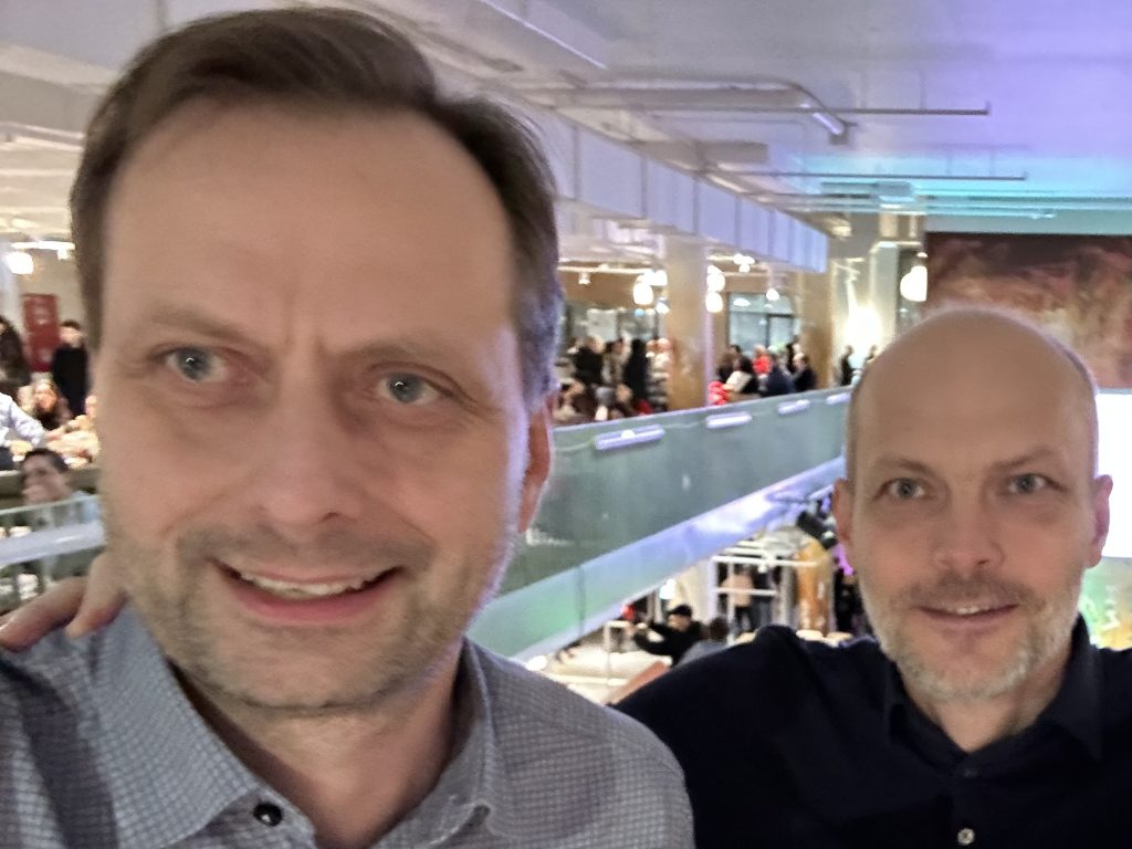 The salfie of two men in their 40s, standing in a big hall